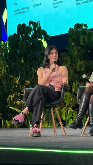 A Babes Net co-founder speaking from a panel chair, microphone in hand, against a wall of greenery