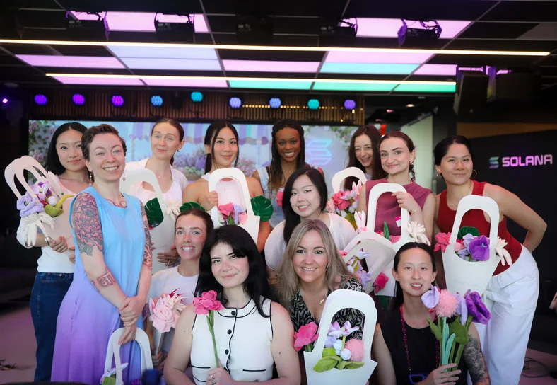 Fourteen women grouped together under pink stage lighting, each holding a white paper bag of flowers