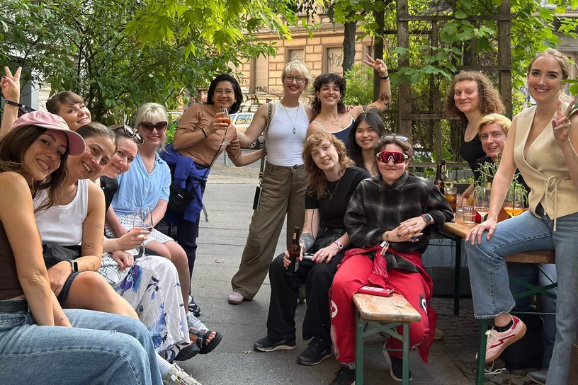 Thirteen women around outdoor tables under the trees, drinks raised to the camera